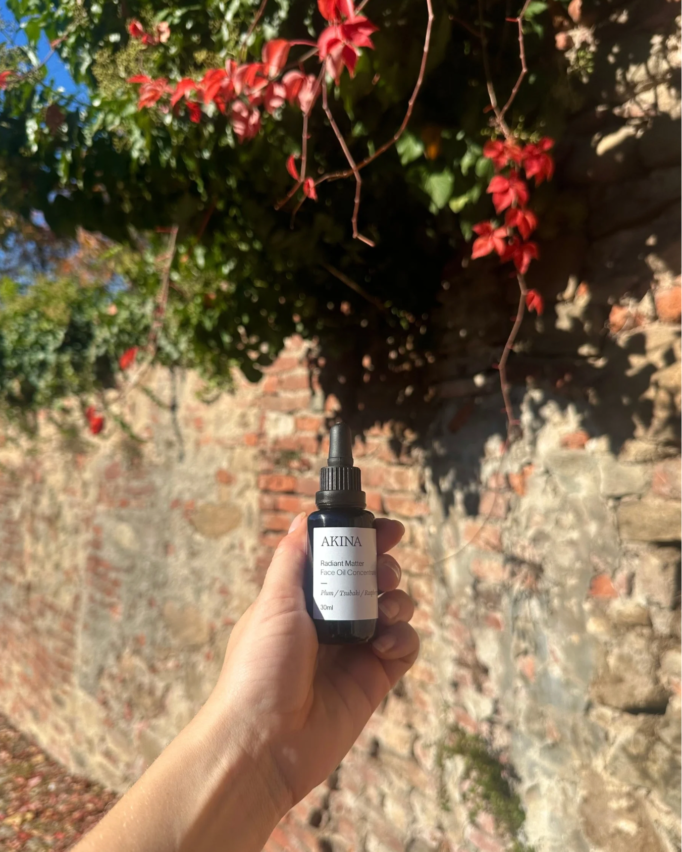 AKINA Radiant Matter face oil held in hand against sunlit leaves and textured stone wall
