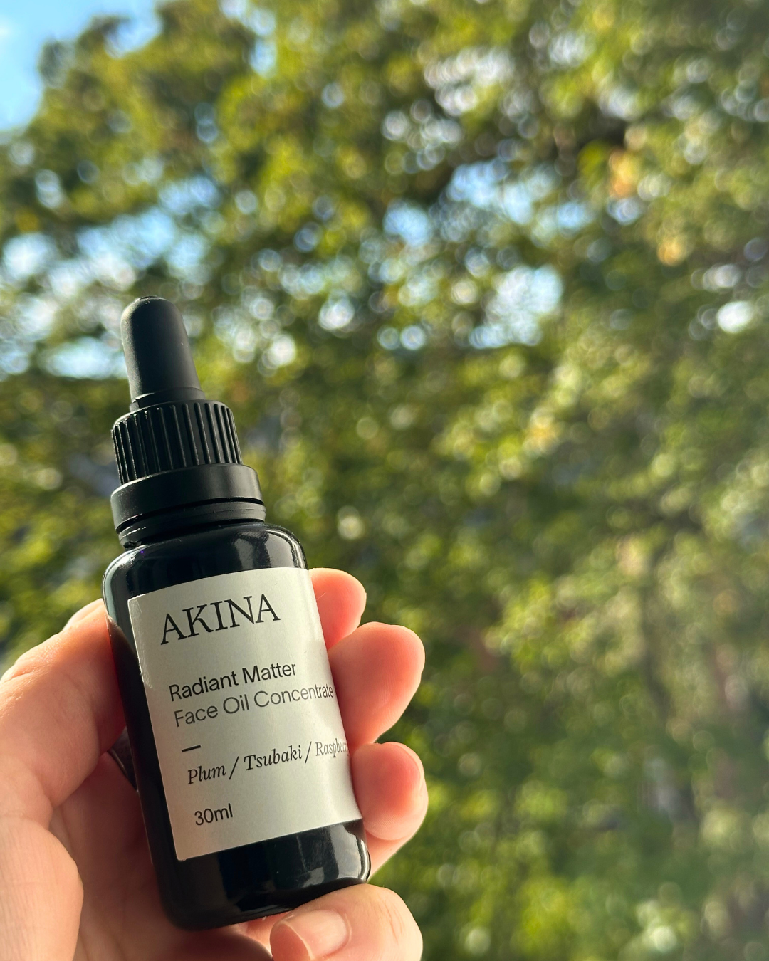 AKINA Radiant Matter face oil held in hand outdoors with green leaves in soft natural light